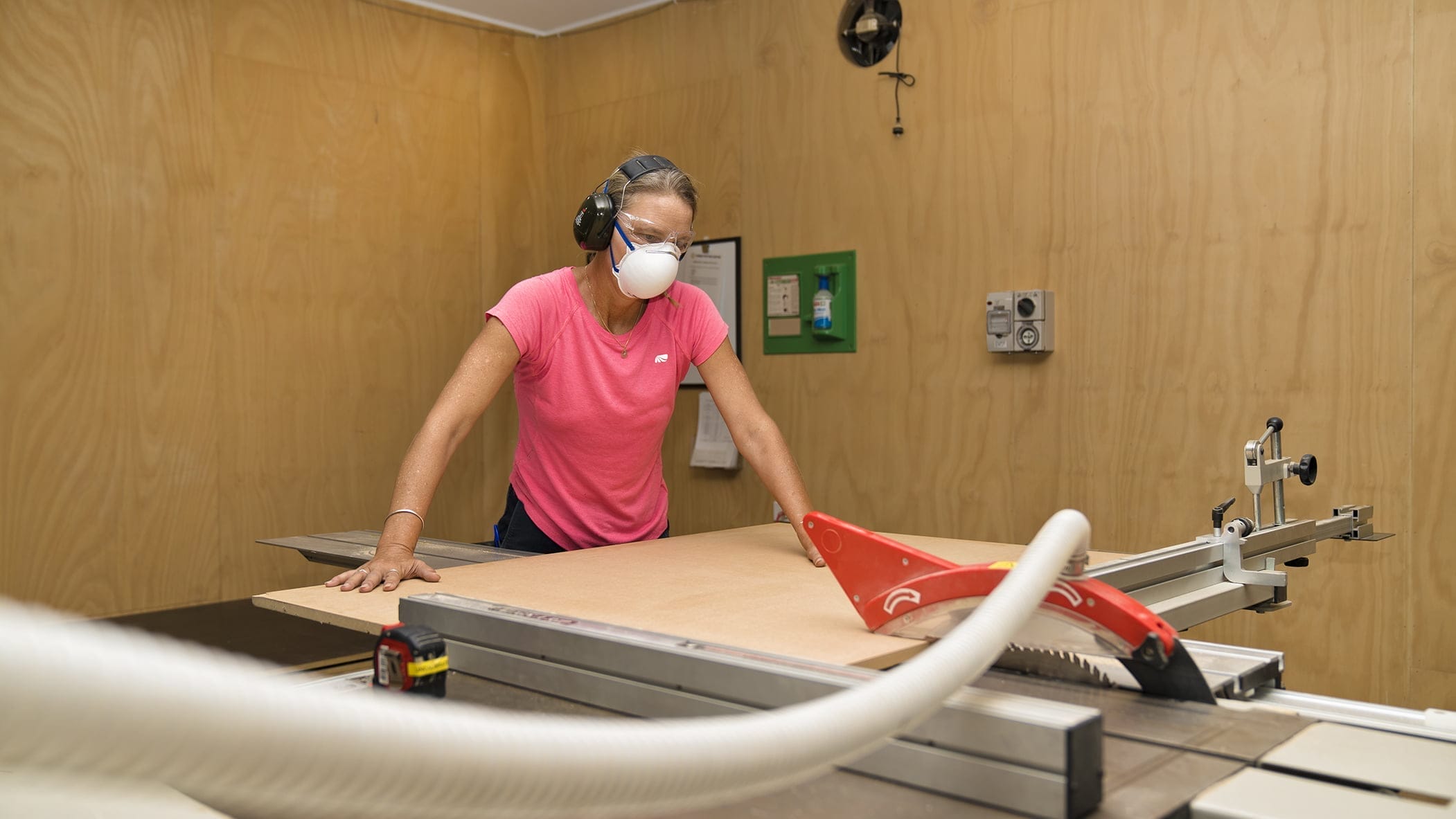 Woman wearing hearing protection, safety glasses and a dust mask pushes a sheet of wood through a table saw with a red blade guard and dust-extraction hose in a plywood-walled workshop.