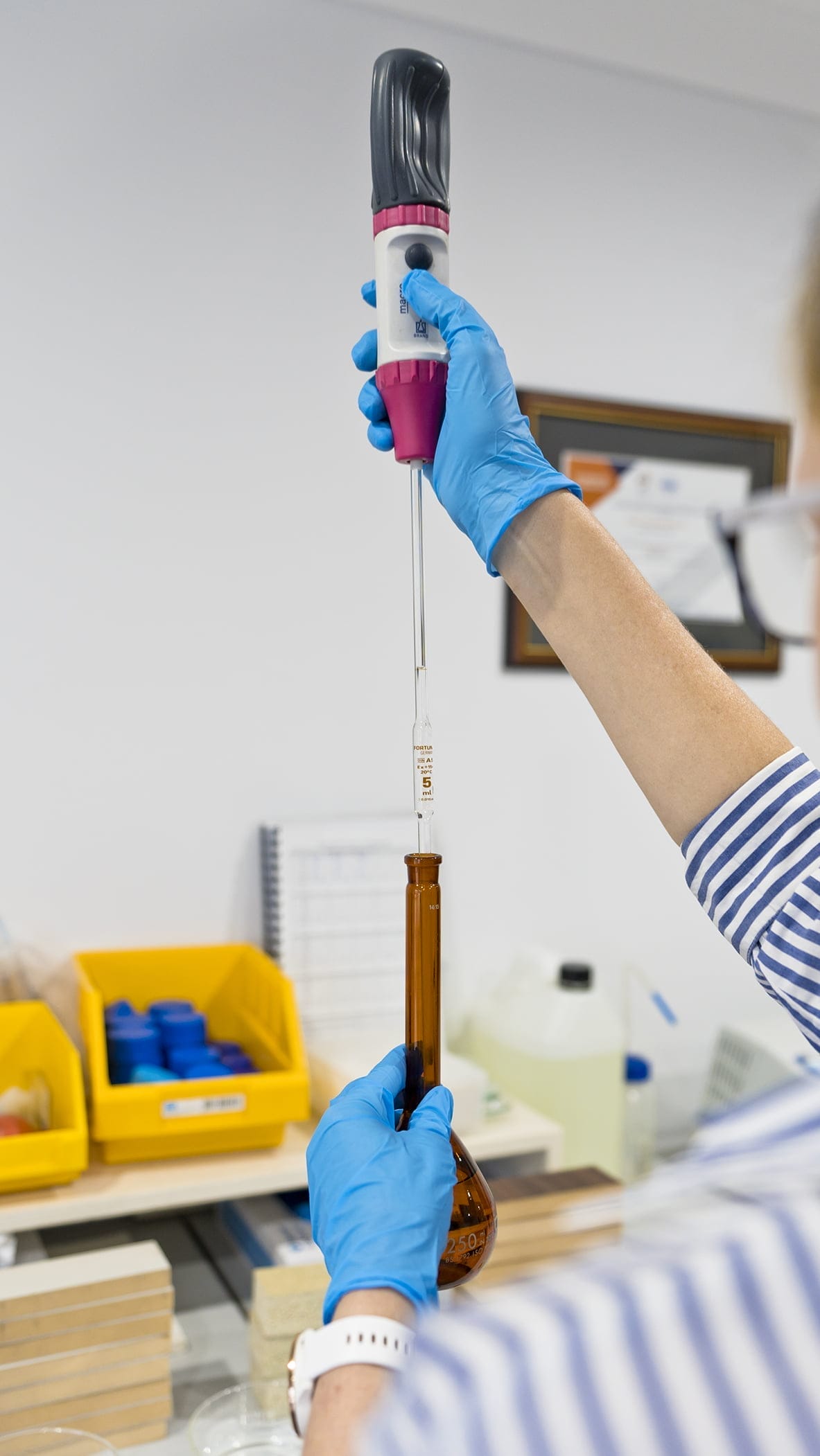 Gloved laboratory worker uses an electronic pipette to dispense liquid through a glass pipette into an amber 250 ml volumetric flask, with yellow storage bins and other bench equipment visible in the background.